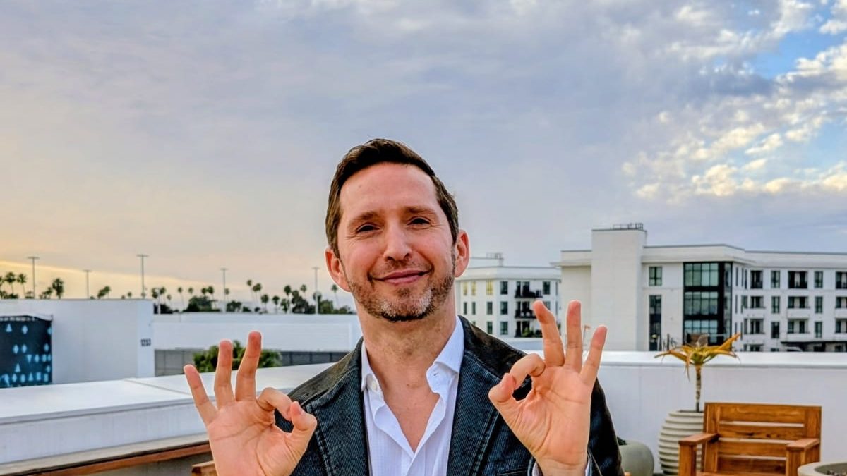 Noel on a rooftop at sunset, smiling, making the OK sign with both hands