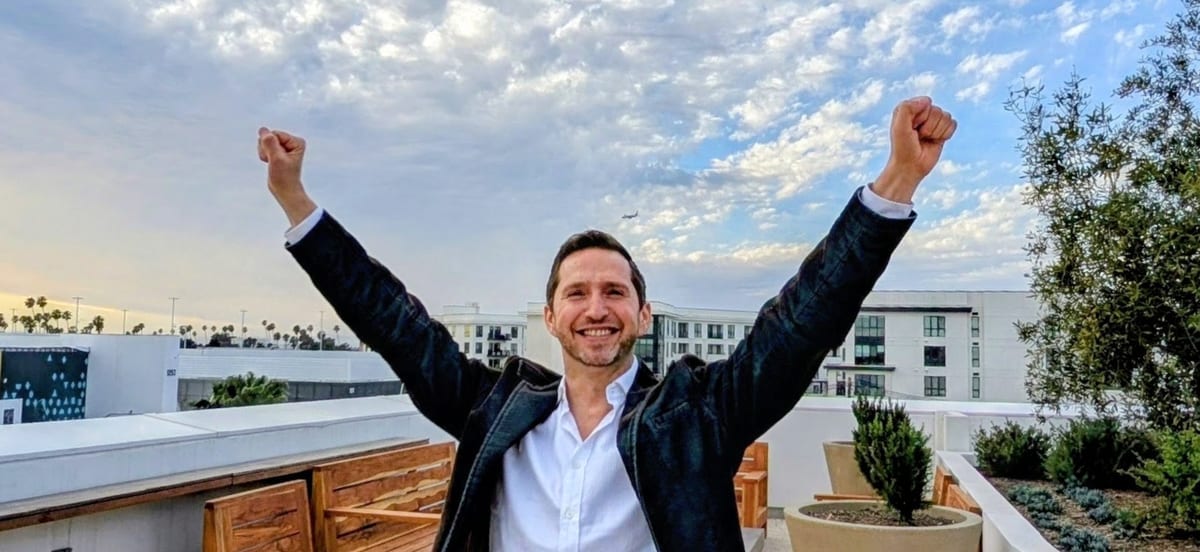 Noel on a rooftop, both arms raised against an open sky
