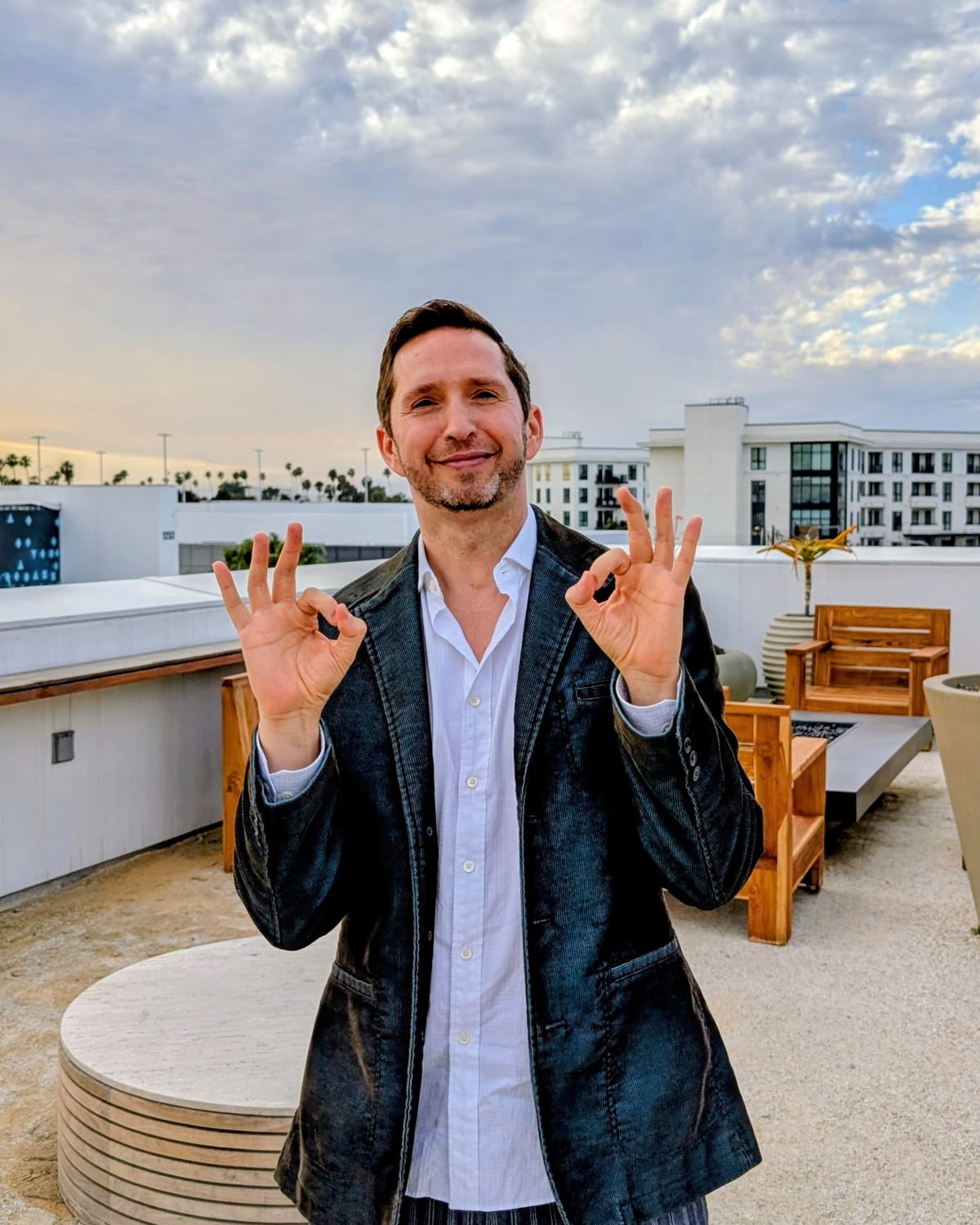 Noel on a rooftop terrace, both hands making an OK sign, smiling