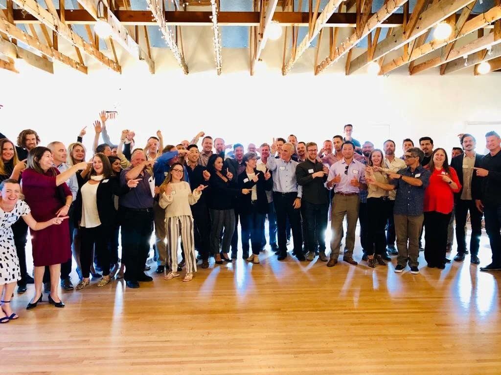 A large workshop group in a bright hall with exposed beams, pointing at the camera at the end of a session