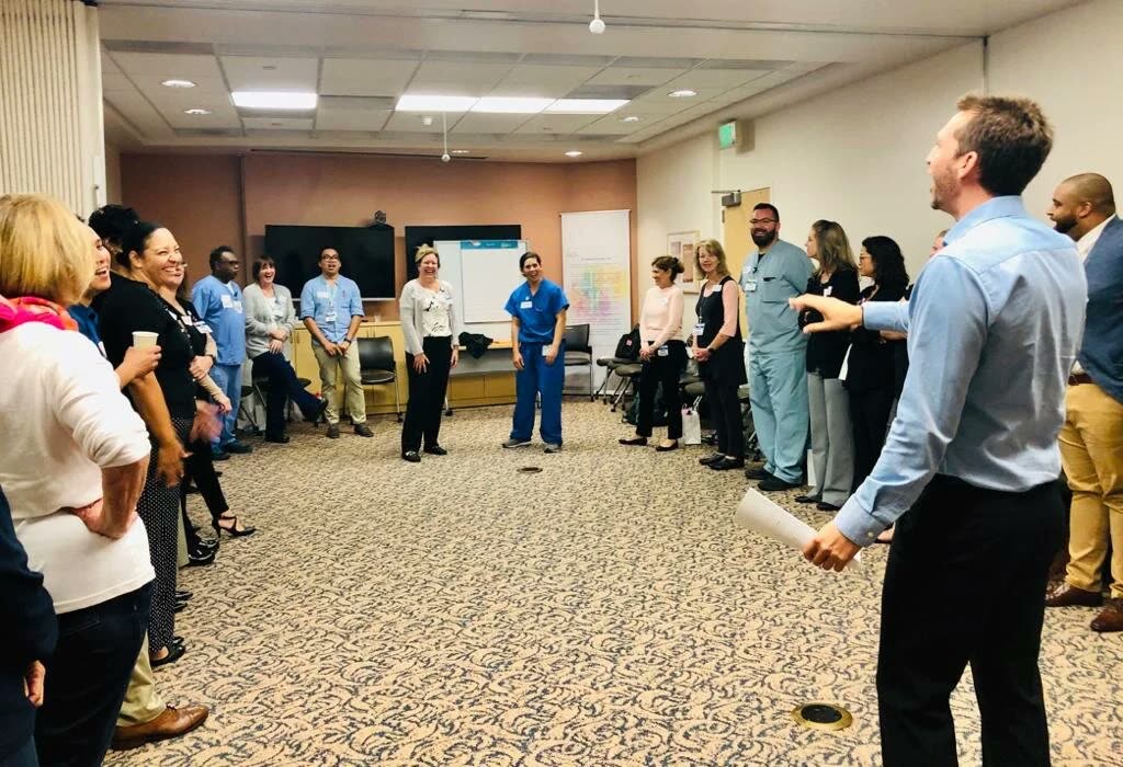 Noel running a session for a hospital team, the room standing in a circle and laughing